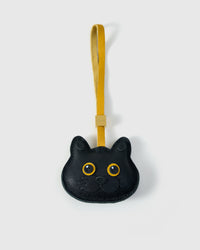 Black British Shorthair cat charm handmade from French Alran genuine goatskin — pure black body, gold eyes, small black nose, mustard-yellow leather strap. Three days by hand. Arrives in luxury gift packaging. Designed in Amsterdam.