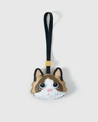 Ragdoll v.1 cat charm handmade from French Alran genuine goatskin — beige body, chocolate-brown markings, soft blue-grey eyes, pink nose, white face mask, black strap. Three days by hand. Arrives in luxury gift packaging. Designed in Amsterdam.
