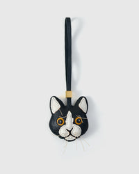 Tuxedo cat charm handmade from French Alran genuine goatskin — black body, white face mask and chest, pink inner ears, gold-yellow eyes, real fiber whiskers, dark navy leather strap. Three days by hand. Arrives in luxury gift packaging. Designed in Amsterdam.