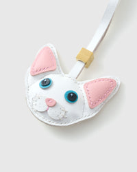 Close-up of the snow-white Devon Rex cat charm — pink inner ears, blue eyes in real leather, pink nose, white face mask. Every panel by hand.