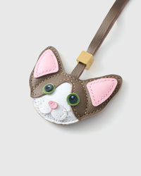 Tight close-up of the taupe Devon Rex face, angled — green eyes in real leather, pink inner ears, white face mask. Every panel by hand.