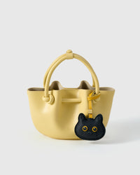Black British Shorthair styled on a pastel-yellow bucket bag, front — a small, specific piece that turns a simple bag into a personal one.