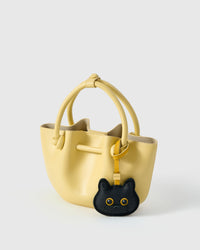 Black British Shorthair styled on a pastel-yellow bucket bag, side angle — real French leather, 3 days by hand.
