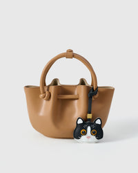 Black Mist British Shorthair styled on a tan bucket bag, front — a small, specific piece that turns a simple bag into a personal one.