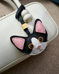 Black-and-white Devon Rex cat charm hanging on a brown suede bag, side angle — real French leather, 3 days by hand.