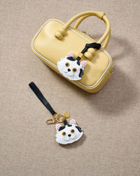 Overhead flatlay — Dragon Li cat charm in strap and keychain editions, alongside a soft-yellow box bag. Finished one at a time, by hand.
