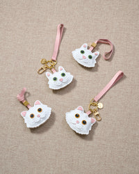 Overhead flatlay — four white Maine Coon charms in strap and keychain editions, arranged in a fan. Finished one at a time, by hand.