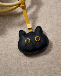 Black British Shorthair charm hanging on a mustard-yellow leather strap, close detail — 5,000+ reviews across STICKYZOO. Real French leather, 3 days by hand.