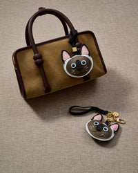 Siamese cat charm styled on a mustard-tan suede top-handle bag, with the keychain edition below. Real French Alran leather, hand-cut over 3 days.