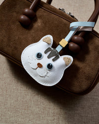 White British Shorthair Cloud Dream charm on a rich brown suede top-handle bag, side angle — rated 4.9★ by thousands of buyers. Real French Alran goatskin, 3 days by hand.