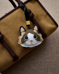 Ragdoll v.1 charm hanging on a tan suede top-handle bag, front angle — a small, specific piece that turns heads.