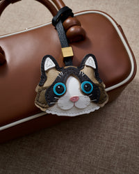 Ragdoll charm hanging on a warm brown leather top-handle bag, side angle — rated 4.9★ by thousands of buyers. Real French Alran leather, 3 days by hand.