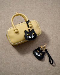 Overhead flatlay — Chubby Midnight British Shorthair in strap and keychain editions, alongside a soft-yellow box bag. Finished one at a time, by hand.