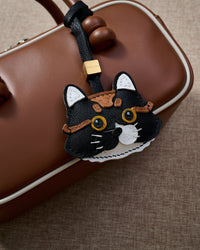 Black long-hair tabby styled on a tan leather top-handle bag, front — a small, specific piece that turns a simple bag into a personal one.