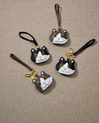 Overhead flatlay — Grey Muse and Black Muse British Shorthair charms grouped on cream surface, in strap and keychain editions. Finished one at a time, by hand.