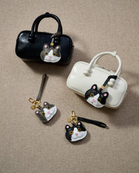 Overhead flatlay — Black Muse and Grey Muse British Shorthair charms in strap and keychain editions, with a black box bag and a cream box bag. Finished one at a time, by hand.