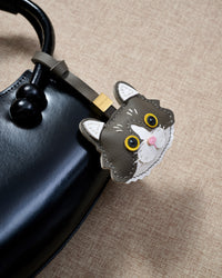 Both editions of the gray European Shorthair side by side — leather strap loop for bags, or 18K gold-plated keychain clip.