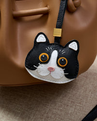 Black Mist British Shorthair hanging on a tan bucket bag, close angle — Real French leather, 3 days by hand.