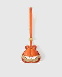 Garfield-style charm handmade from French Alran genuine goatskin — orange tabby face with darker stitched stripes, half-closed eyes, sandy muzzle with small frown, orange leather strap. Three days by hand. Arrives in luxury gift packaging. Designed in Amsterdam.