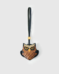 Owl charm handmade from French Alran genuine goatskin — copper-and-tan layered scalloped feathers, cream eyebrow, bright yellow eyes with dark pupils, yellow beak, black leather strap. Three days by hand. Arrives in luxury gift packaging. Designed in Amsterdam.
