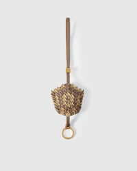 Scaled pinecone charm handmade from French Alran genuine goatskin — dozens of hand-cut scales in gold and taupe-gray, layered into a dimensional form, taupe leather strap with gold ring. Five days by hand. Arrives in luxury gift packaging. Designed in Amsterdam.