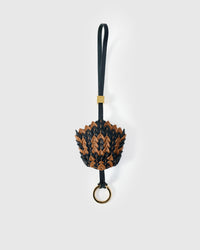 Scaled pinecone charm handmade from French Alran genuine goatskin — dozens of hand-cut scales in black and caramel, layered into a dimensional form, black leather strap with gold ring. Five days by hand. Arrives in luxury gift packaging. Designed in Amsterdam.