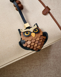 Close-up of the owl charm on a cream canvas bag — every scalloped feather layer cut and hand-stitched in real French leather, yellow eyes in real leather, yellow beak.