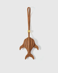 Sculpted caramel whale charm handmade from French Alran genuine goatskin — dimensional 3D body viewed from above, side fins, split tail fluke, cream hand-stitched seams, tan leather strap. Three days by hand. Arrives in luxury gift packaging. Designed in Amsterdam.