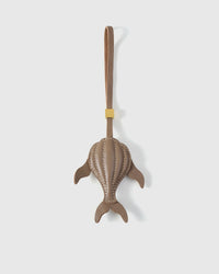Sculpted taupe whale charm handmade from French Alran genuine goatskin — dimensional 3D body viewed from above, side fins, split tail fluke, cream hand-stitched seams, taupe leather strap. Three days by hand. Arrives in luxury gift packaging. Designed in Amsterdam.