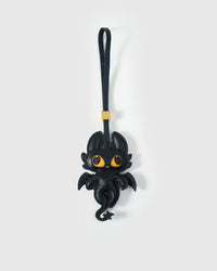 Black dragon charm handmade from French Alran genuine goatskin — deep black body, bat-like wings, large amber-yellow eyes, curled leather tail, black leather strap. Three days by hand. Arrives in luxury gift packaging. Designed in Amsterdam.