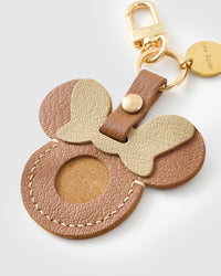 Close-up of the tan Mickey AirTag holder — gold-cream bow, stitched edges, warm tan goatskin. Every panel by hand.