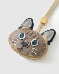 Tight close-up of the Tonkinese face, angled — pale blue eyes in real leather, dark forehead stripes, tan ear centres, black nose. Every panel by hand.