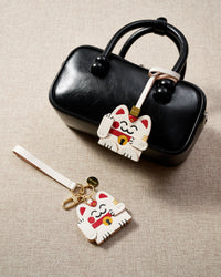 Both editions of the Maneki Neko (lucky cat) charm side by side — leather strap loop for bags, or 18K gold-plated keychain clip with engraved STICKYZOO disc.