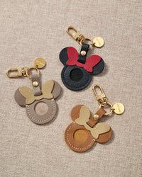 Close-up of the taupe Mickey AirTag holder — gold bow, stitched edges, cool taupe goatskin. Every panel by hand.