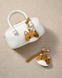 Overhead flatlay — Persian cat charm in strap and keychain editions alongside a cream top-handle bag. Finished one at a time, by hand.