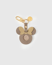 Taupe Mickey-silhouette AirTag holder handmade from French Alran genuine goatskin — cool taupe-grey body, gold bow, round Mickey ears, precision AirTag cavity, 18K gold-plated keychain clip. Three days by hand. Arrives in luxury gift packaging. Designed in Amsterdam.