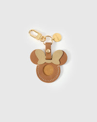 Tan Mickey-silhouette AirTag holder handmade from French Alran genuine goatskin — warm tan body, gold-cream bow, round Mickey ears, precision AirTag cavity, 18K gold-plated keychain clip. Three days by hand. Arrives in luxury gift packaging. Designed in Amsterdam.
