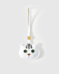 Green-eyes Scottish Fold cat charm handmade from French Alran genuine goatskin — pure white body, grey forehead stripes, vivid green eyes, copper nose, white leather strap. Three days by hand. Arrives in luxury gift packaging. Designed in Amsterdam.