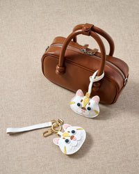 Overhead — Light Silver Tabby in strap and keychain editions alongside a warm brown leather box bag. Finished one at a time, by hand.