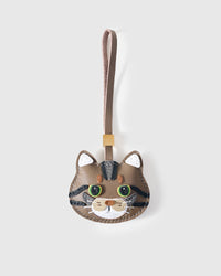 Tabby cat charm handmade from French Alran genuine goatskin — warm greige body, dark tabby stripes, green eyes, white ear tufts, cream muzzle, taupe leather strap. Three days by hand. Arrives in luxury gift packaging. Designed in Amsterdam.