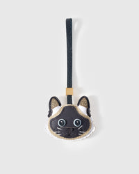 Siamese cat charm handmade from French Alran genuine goatskin — cream body, chocolate-black colour-point mask, pale blue eyes, tan-centred ears, black leather strap. Three days by hand. Arrives in luxury gift packaging. Designed in Amsterdam.