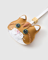 Angled close-up — every stitch visible across the ginger British Shorthair face. Real French leather, layered panel by panel.