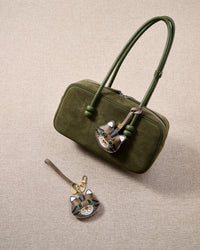Overhead flatlay — tabby cat in strap and keychain editions alongside an olive-green suede bag. Finished one at a time, by hand.