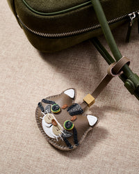Tabby cat charm close on an olive-green suede crossbody, side angle — real French leather, 3 days by hand.