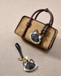 Overhead flatlay — Siamese cat in strap and keychain editions alongside a tan suede top-handle bag. Finished one at a time, by hand.