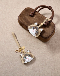 Overhead flatlay — Latte Paws Ragdoll in strap and keychain editions, alongside a brown suede box bag. Finished one at a time, by hand.