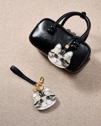 Overhead flatlay — Silver Tabby American Shorthair in strap and keychain editions alongside a black top-handle bag. Finished one at a time, by hand.