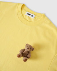 Angled close-up of plush teddy bear on lemon cotton showing 3D texture with neckline and label visible