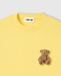 Close-up of full-body plush teddy bear with brown fur, stitched paws, and embroidered face on lemon cotton