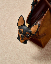 Handmade Chihuahua leather bag charm clipped to a brown suede handbag — French Alran leather and suede, 18K gold-plated hardware. STICKYZOO collectible.